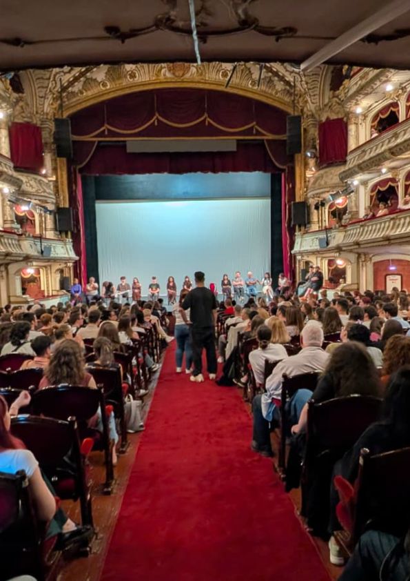 Audience fills a ornate theater, facing a stage where a line of performers stands beneath a blue backdrop on a red-carpet aisle. Balcony boxes with gold detailing surround the scene.