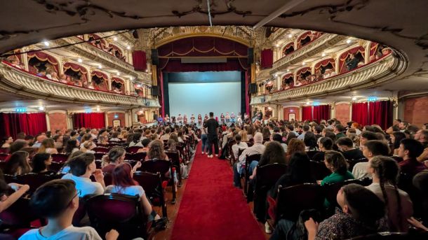 Audience fills a ornate theater, facing a stage where a line of performers stands beneath a blue backdrop on a red-carpet aisle. Balcony boxes with gold detailing surround the scene.