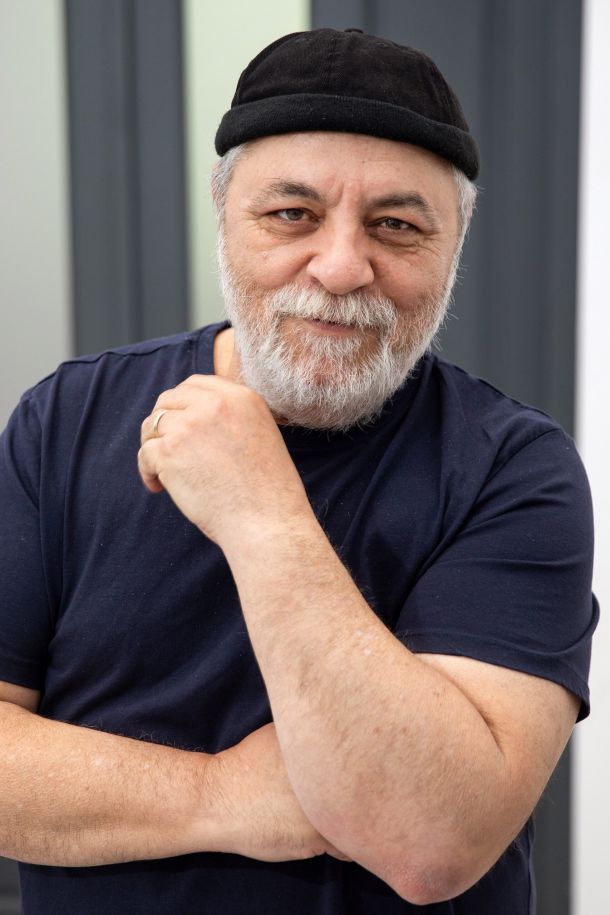 Older man with a gray beard wearing a black cap and navy t-shirt, arms crossed and a subtle smile.